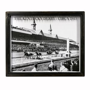 May include: A black and white photograph of the Kentucky Derby, circa 1943. The photo shows a view of the finish line with horses and jockeys racing towards the finish line. The photo is framed in a black frame.