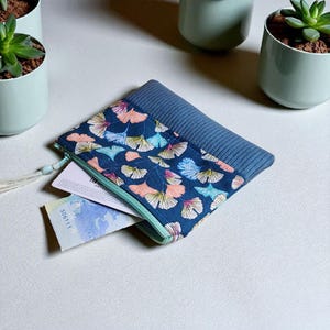 Peut inclure: Une pochette bleue zippée avec un motif floral de feuilles roses, jaunes et bleues sur fond bleu marine. La partie supérieure est en tissu côtelé bleu uni. La pochette est ouverte, révélant quelques cartes.