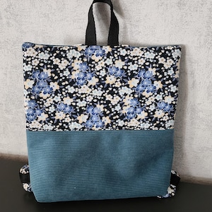 Peut inclure: Un sac à dos floral avec une poignée et des bretelles noires. La partie supérieure présente un fond noir avec des fleurs bleues et blanches. La partie inférieure est en velours côtelé bleu sarcelle. Le sac est conçu pour transporter des objets.