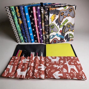 Peut inclure: Ensemble de cahiers recouverts de tissu et un étui à stylos enroulable. Les cahiers présentent divers motifs, dont des étoiles, des motifs géométriques et une impression de manette de jeu vidéo. L'étui à stylos est rouge avec un motif de cerf et de feuilles blanches.