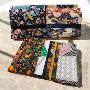 Peut inclure: Trois pochettes en tissu aux motifs floraux et tropicaux. Deux sont pliées, tandis qu'une est ouverte, révélant une fermeture éclair jaune, une carte blanche et une poche en filet contenant plusieurs pilules circulaires blanches et roses.