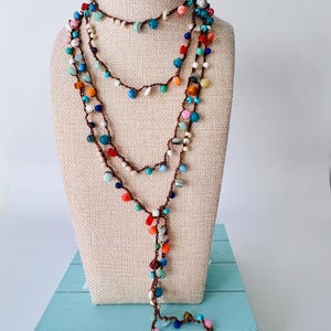May include: A long, multi-strand necklace featuring a brown crocheted cord interspersed with a variety of small, colorful beads in shades of blue, red, orange, pink, and white. The necklace is arranged in multiple loops with a dangling end section.