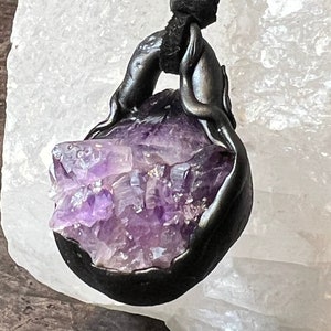 May include: A purple amethyst crystal pendant encased in a black metal setting, hanging from a black leather cord.