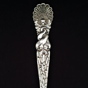 May include: A silver spoon with an ornate design featuring a stylized bird and floral motifs. The handle is shaped like a fan and the bowl is rounded.
