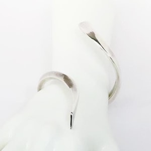 May include: Two silver bangle bracelets on a white mannequin hand. The bracelets are both open and have a curved, organic shape.