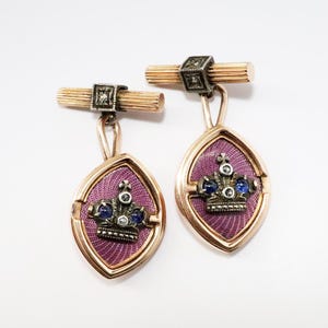 May include: A pair of gold cufflinks with a purple enamel design featuring a crown with blue and white gemstones.