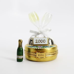 May include: A gold and white porcelain trinket box with a champagne bottle and two champagne glasses on top. The box is decorated with a floral pattern and the year 2000 is inscribed on the front and bottom.