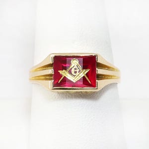May include: A gold-colored ring with a rectangular, ruby-red stone. The stone features a gold-colored Masonic symbol. The ring is displayed on a white surface.