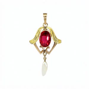 May include: A gold pendant featuring an oval, deep red gemstone at the center. The pendant has ornate gold filigree details and a teardrop-shaped pearl hanging below. The design includes stylized wings and geometric shapes.