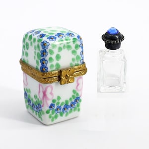 May include: A small, square porcelain trinket box with a hinged lid, decorated with blue flowers, green leaves, and pink bows. A small, clear glass bottle with a blue gem-topped stopper is next to the box.