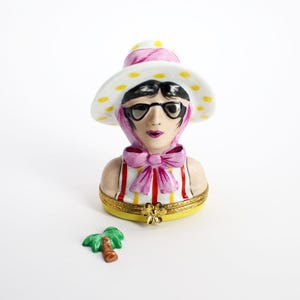 May include: A porcelain figurine of a woman wearing a white hat with yellow polka dots, pink sunglasses, and a pink and white striped dress. The figurine is holding a small palm tree.