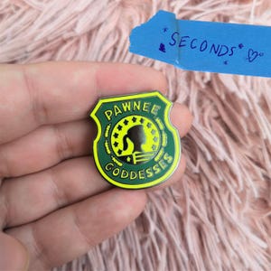 May include: A green and yellow enamel pin featuring the words "Pawnee Goddesses" and a silhouette of a woman's profile. The pin is shield-shaped. A blue piece of tape with the word "SECONDS" is in the background.