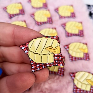 May include: A yellow enamel pin shaped like a wheel of brie cheese with grill marks, a golden wedge, and a red and white checkered napkin. The pin is held in a hand, with several similar pins in the background.