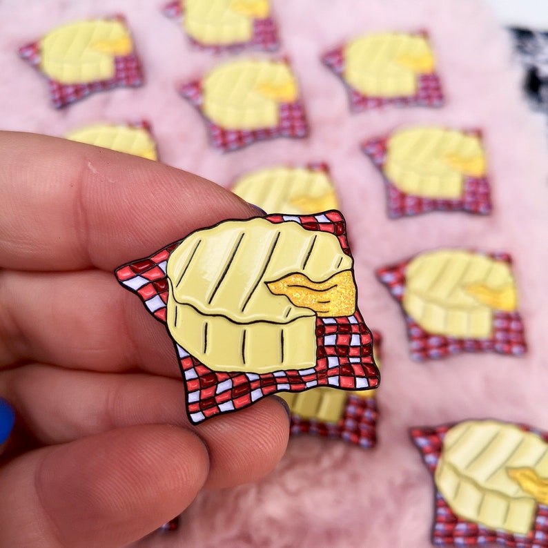 May include: A yellow enamel pin shaped like a wheel of brie cheese with grill marks, a golden wedge, and a red and white checkered napkin. The pin is held in a hand, with several similar pins in the background.