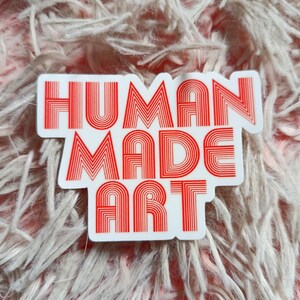 May include: A white sticker with the words "HUMAN MADE ART" in red, retro-style lettering. The sticker is rectangular with rounded corners and is set against a soft, pinkish-white, fuzzy background.