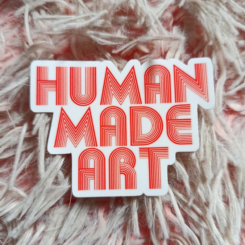 May include: A white sticker with the words "HUMAN MADE ART" in red, retro-style lettering. The sticker is rectangular with rounded corners and is set against a soft, pinkish-white, fuzzy background.