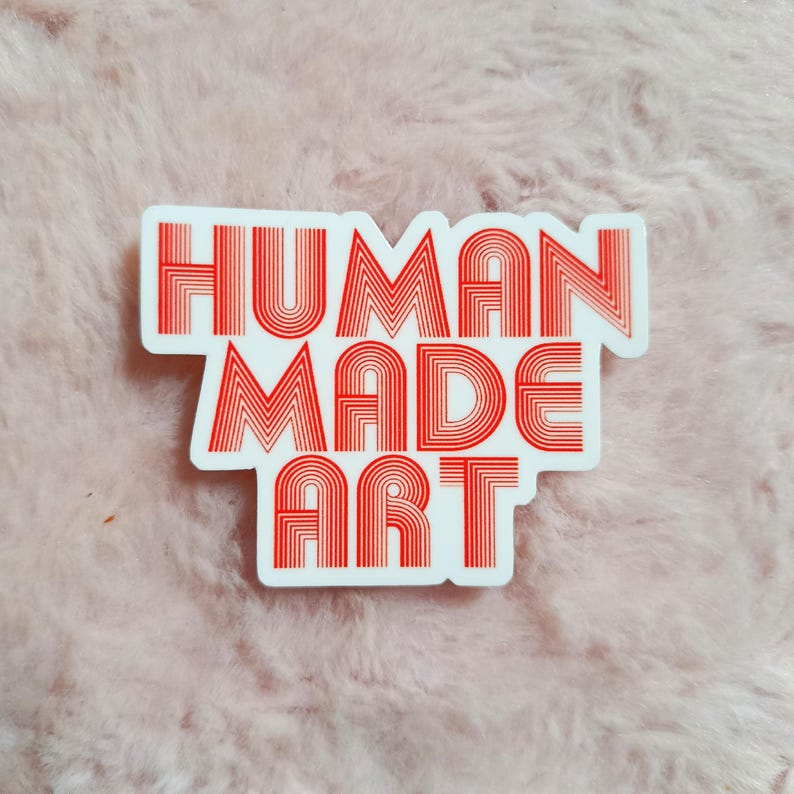May include: A white sticker with the words "HUMAN MADE ART" in red, stylized, layered font. The sticker has a rectangular shape with rounded corners. The background is a soft, pink, textured surface.