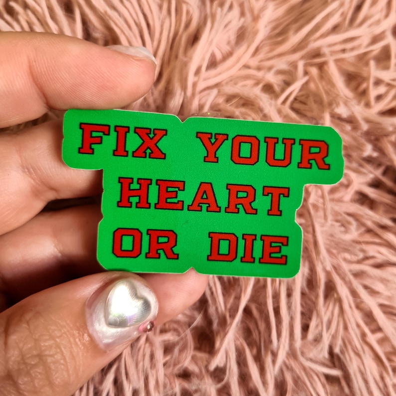 May include: A green sticker with the text "FIX YOUR HEART OR DIE" in red block letters. The sticker is held in a hand against a pink, fuzzy background.