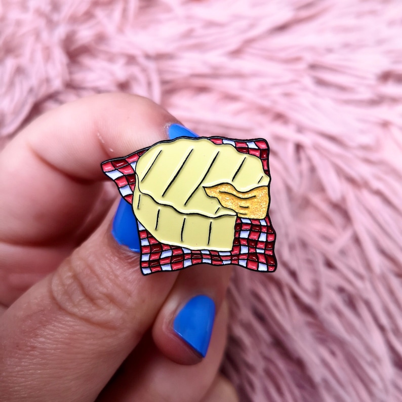 May include: A yellow and black enamel pin shaped like a grilled cheese sandwich on a red and white checkered napkin. The pin has a bite taken out of it, revealing a golden interior.