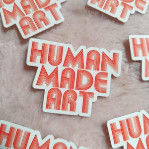 May include: White stickers with the words "HUMAN MADE ART" in red, retro-style lettering. The stickers are scattered on a pink, fuzzy surface. The text is in a bold, sans-serif font.