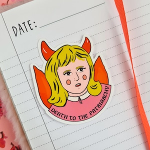 May include: A sticker featuring a cartoon illustration of a person with blonde hair, red horns, and orange wings. The person is wearing a pink dress, and the sticker has the text "DEATH TO THE PATRIARCHY". The sticker is on a lined notebook.