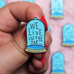 Pode incluir: Um broche de esmalte azul com uma cúpula de vidro e o texto "WE LIVE IN THE JAR - SYLVIA PLATH" dentro.