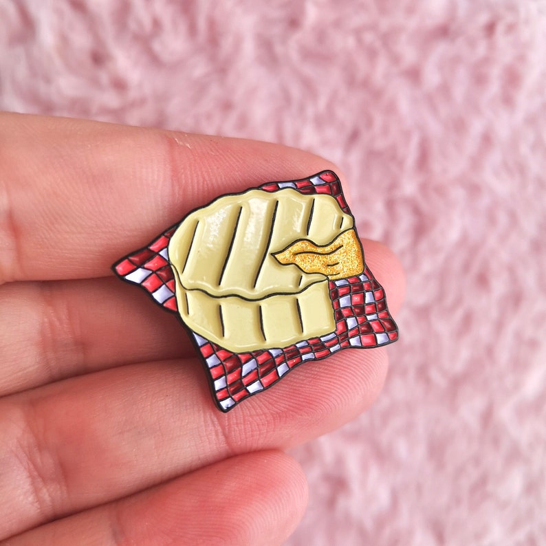 May include: A cheese enamel pin featuring a grilled cheese design with a golden-yellow color, resting on a red and white checkered napkin. The pin has a black outline and a small, glittery gold accent.