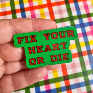 May include: A green sticker with the text "FIX YOUR HEART OR DIE" in red block letters. The sticker is held against a colorful plaid background.