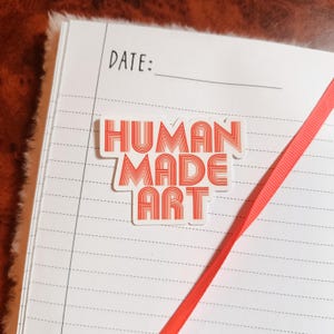 May include: A white notebook with lined pages and a red ribbon bookmark. A sticker with the words "HUMAN MADE ART" in red lettering is affixed to the notebook. The word "DATE:" is printed at the top.