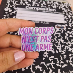 Può includere: Un adesivo olografico con il testo "MON CORPS N'EST PAS UNE ARME" in lettere rosa e viola su sfondo bianco.