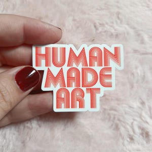 May include: A white sticker with the words "HUMAN MADE ART" in red, retro-style lettering. The sticker is held against a soft, pink background.