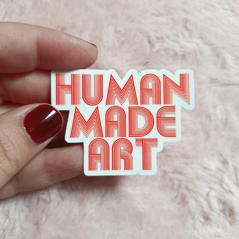 May include: A white sticker with the words "HUMAN MADE ART" in red, retro-style lettering. The sticker is held against a soft, pink background.