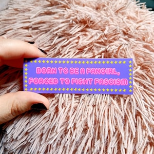 May include: A rectangular sticker with the text "BORN TO BE A FANGIRL, FORCED TO FIGHT FASCISM" in pink and yellow on a purple background. The sticker is bordered with yellow stars. The sticker is held by a hand with black nail polish.