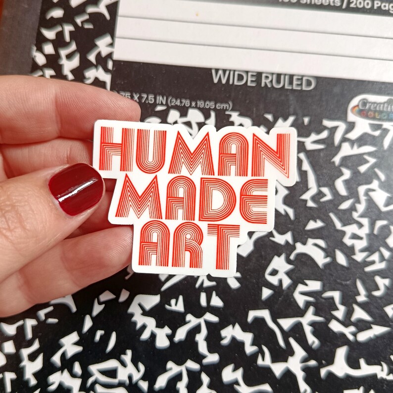 May include: A white sticker with the words "HUMAN MADE ART" in red, stylized lettering. The sticker is rectangular and held against a black and white patterned background. The sticker measures 7.5 inches (19.05 cm) in length.