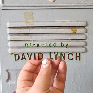 May include: A clear sticker with the text "Directed by DAVID LYNCH" in green and red lettering is held in front of a gray metal surface. The sticker is held by a hand with a heart-shaped nail design.