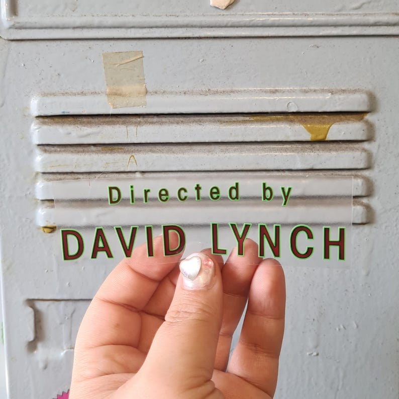 May include: A clear sticker with the text "Directed by DAVID LYNCH" in green and red lettering is held in front of a gray metal surface. The sticker is held by a hand with a heart-shaped nail design.