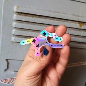 May include: A holographic sticker featuring a constellation with black stars and a dashed line outline. The sticker is iridescent with shades of blue, pink, and yellow.