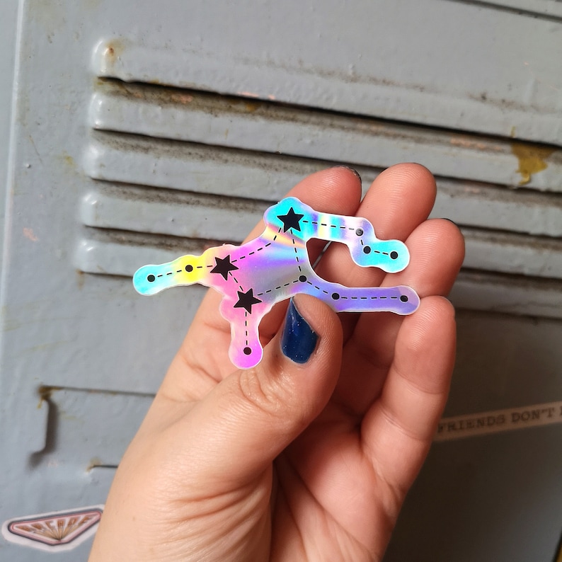 May include: A holographic sticker featuring a constellation with black stars and a dashed line outline. The sticker is iridescent with shades of blue, pink, and yellow.
