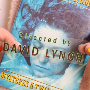 May include: A book cover with a blue and white image of a person's face. The text "Directed by DAVID LYNCH" is overlaid on a clear sticker. The title "Myst&egrave;res a Twin Peaks" is visible at the bottom.