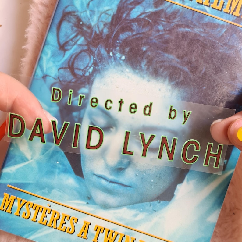 May include: A book cover with a blue and white image of a person's face. The text "Directed by DAVID LYNCH" is overlaid on a clear sticker. The title "Myst&egrave;res a Twin Peaks" is visible at the bottom.