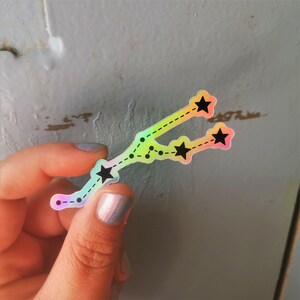 May include: A holographic sticker featuring a constellation of stars in a rainbow colour scheme. The sticker is shaped like the constellation Taurus, with five stars and a dotted line connecting them.
