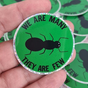 Puede incluir: Una pegatina redonda verde y negra con un borde de purpurina plateado. La pegatina presenta una hormiga negra con el texto "We are many they are few".