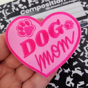 Peut inclure: Patch en forme de cœur rose avec le texte "DOG MOM" en lettres roses. Deux empreintes de pattes roses sont de chaque côté du texte.