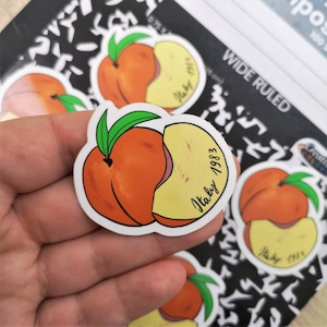 May include: A sticker of a peach with a bite taken out of it. The peach is orange and yellow with a green leaf. The sticker says "Italy 1983" in black cursive writing.