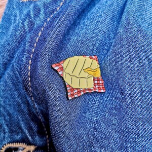 May include: A gold-colored enamel pin shaped like a pie, resting on a red and white checkered napkin. The pie has a golden crust and a filling that is partially visible. The pin is set against a blue denim background.