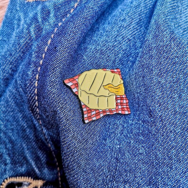 May include: A gold-colored enamel pin shaped like a pie, resting on a red and white checkered napkin. The pie has a golden crust and a filling that is partially visible. The pin is set against a blue denim background.