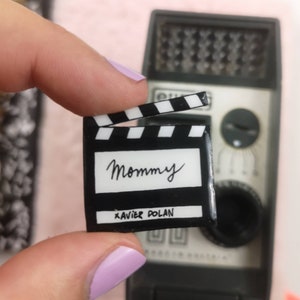 Peut inclure: Une claquette noire et blanche avec le mot "Mommy" écrit en cursive et le nom "Xavier Dolan" imprimé en dessous.