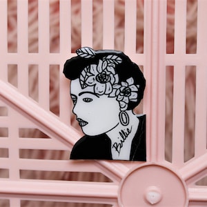 Puede incluir: Pin de esmalte negro y blanco con el retrato de una mujer con flores en el pelo. La mujer tiene un tatuaje que dice "Billie" en el cuello.
