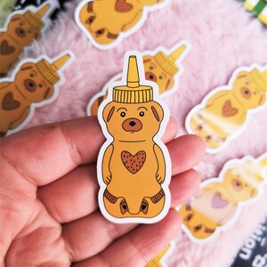 Peut inclure: Un ensemble d'autocollants en forme de bouteille de miel d'ours jaunes. Les autocollants représentent un ours de dessin animé avec un cœur sur la poitrine.