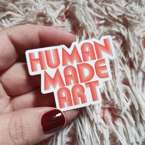 May include: A rectangular sticker with the words "HUMAN MADE ART" in red, retro-style lettering. The sticker is white with a red border and is held by a hand with red nail polish.